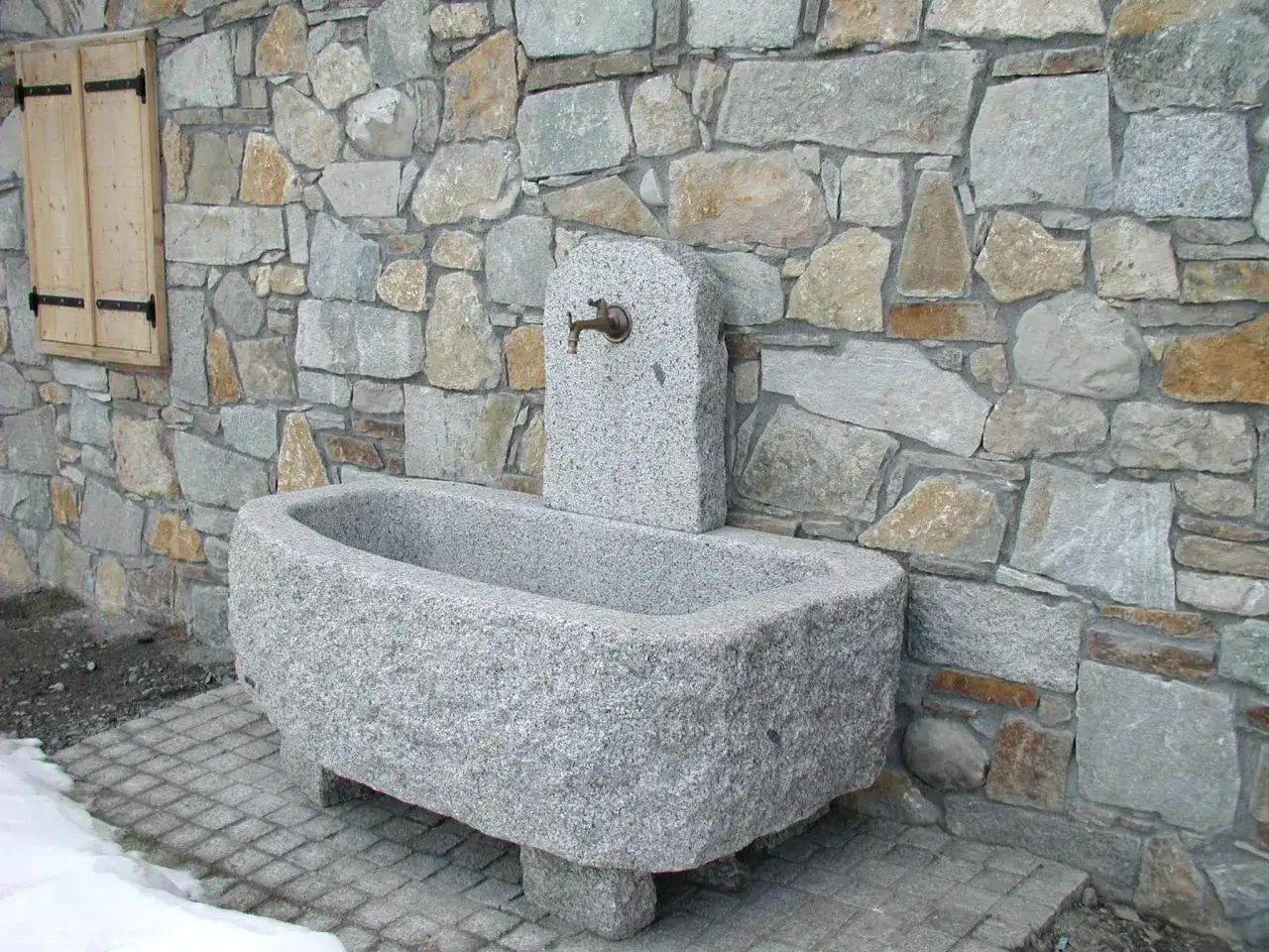 Fontaine en pierre sculptée, avec une bordure de bassin en pierre rustique, adossée à un mur de pierres sèches.