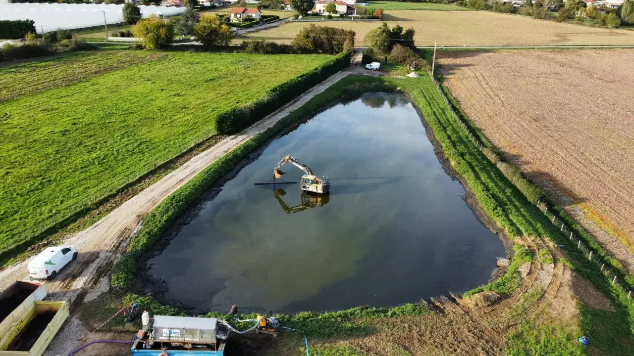 Un excavateur travaille dans un étang, entouré de champs verts et de roseaux. Une pompe est installée sur la rive.